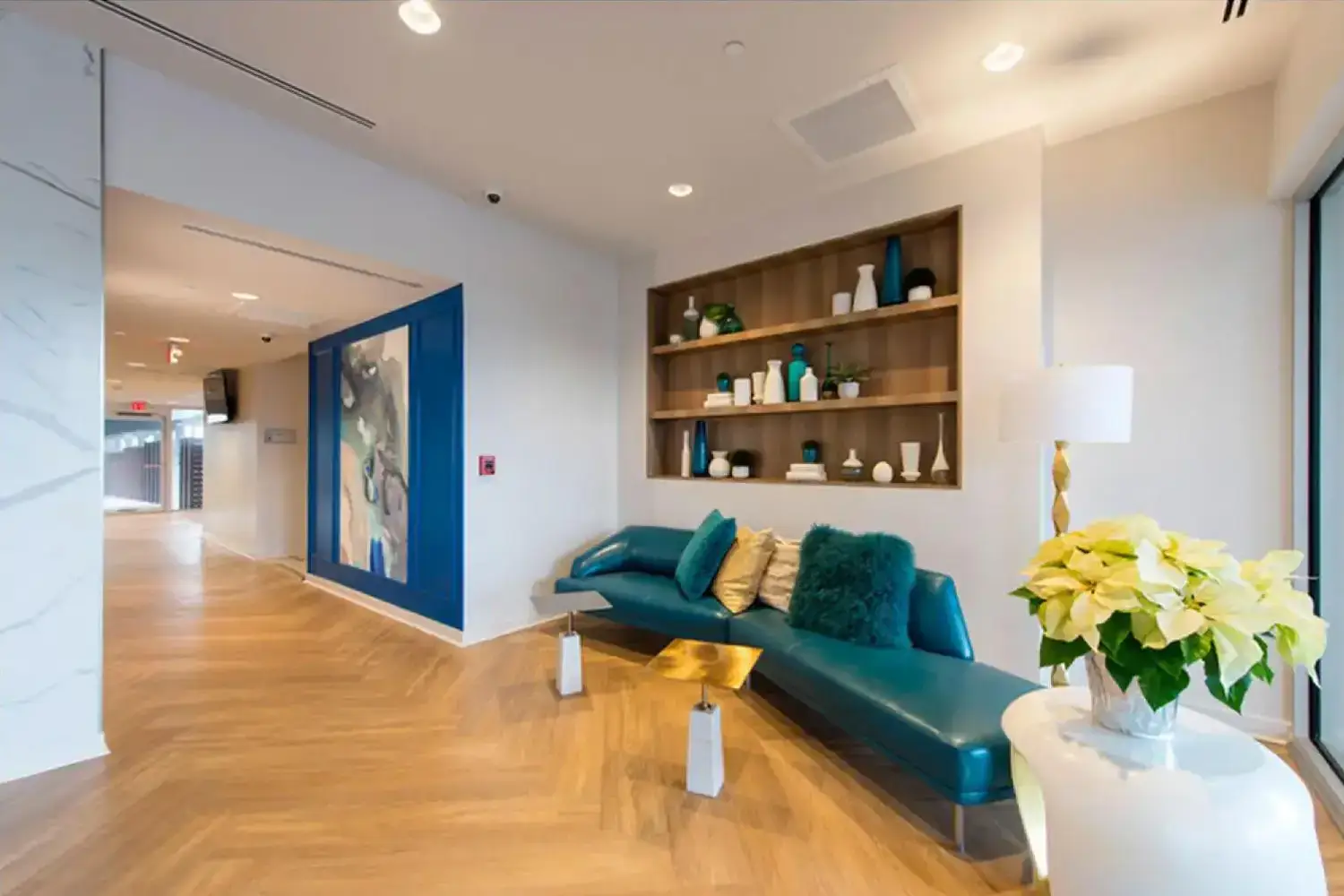 Modern and luxurious waiting area with a blue leather couch, green and gold throw pillows, white coffee table, and a hallway with a wooden floor.