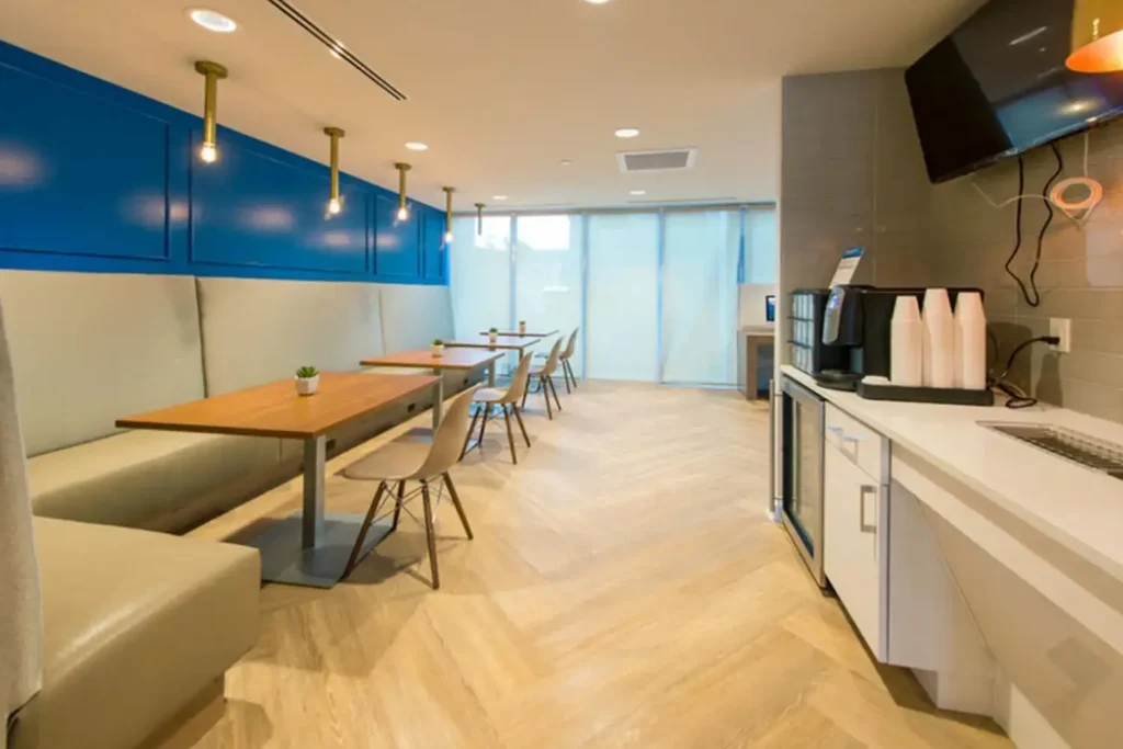 Bright community Coffee Lounge and dining area with gray countertop, wooden table, blue chairs, and blue pendant lights.