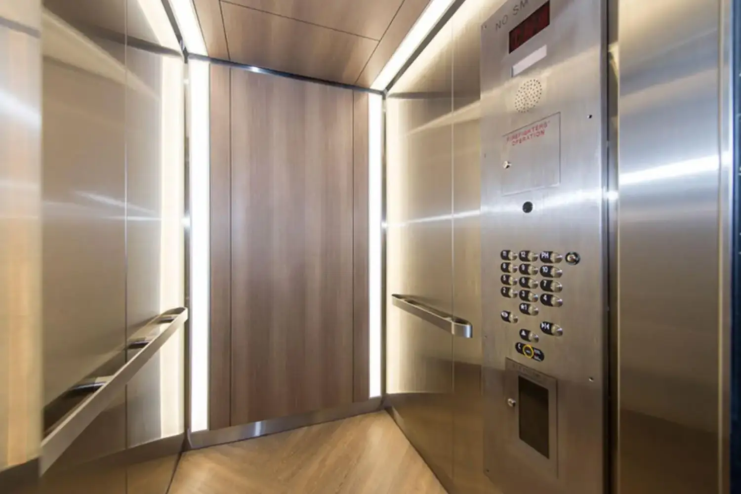 Interior of an elevator with wooden paneling, mirrored ceiling, control panel, handrail, and a NO SMOKING sign.