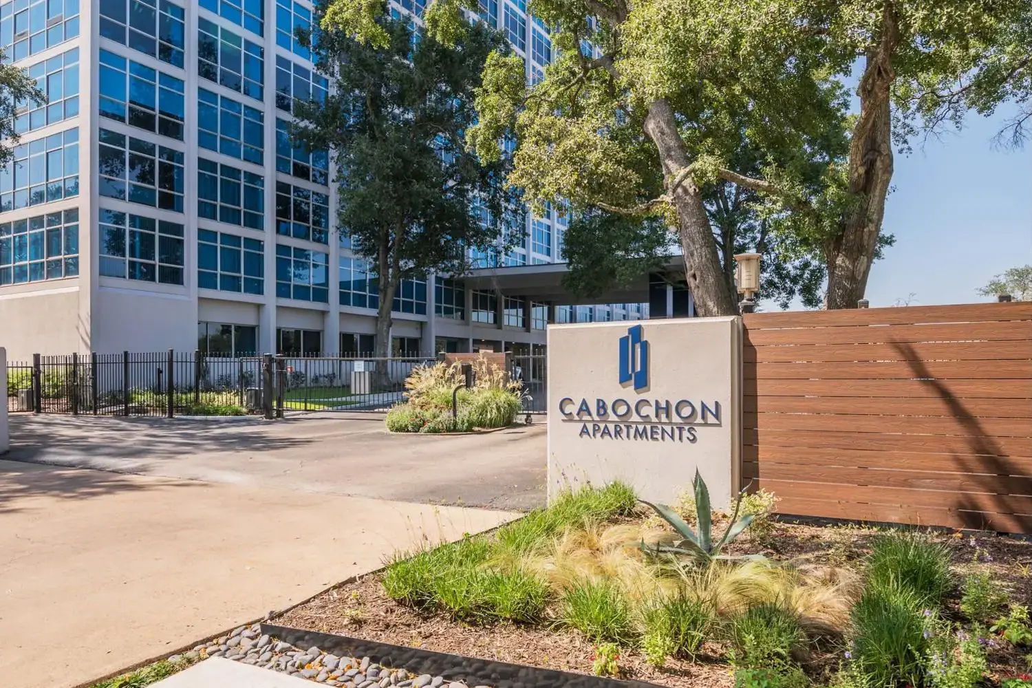 Entrance of Cabochon Apartments, a modern high-rise building with blue-tinted windows, landscaped with trees, shrubs, and grasses.
