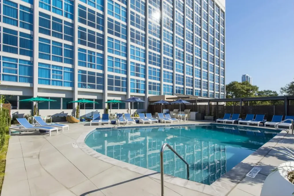 Large outdoor swimming pool in front of a modern high-rise building with blue-tinted windows, surrounded by lounge chairs.