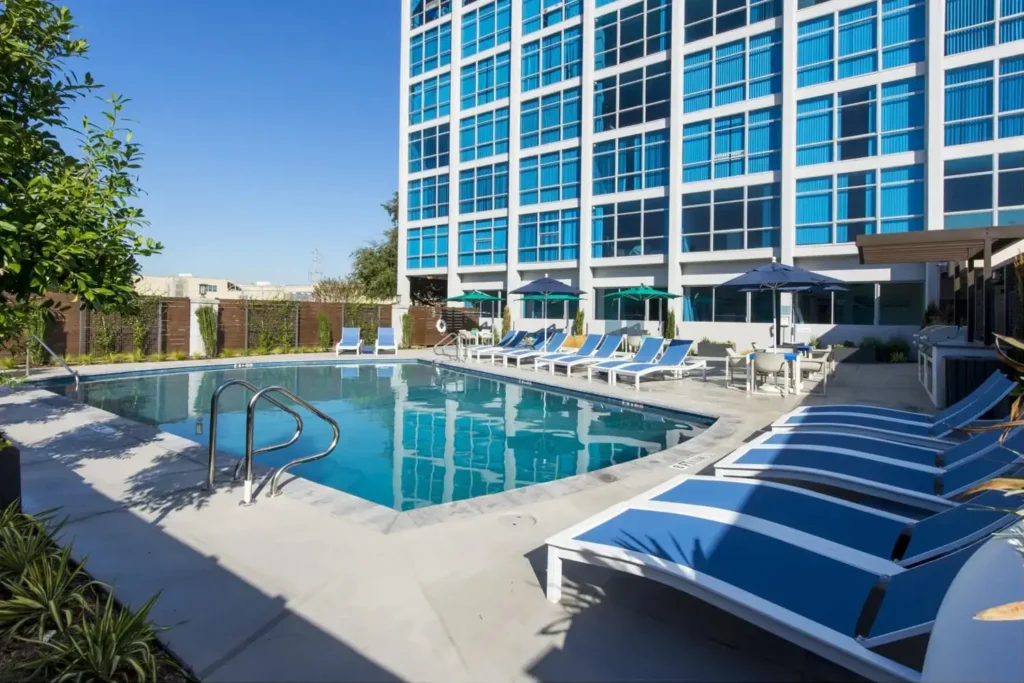 Community area with a rectangular swimming pool, white & blue lounge chairs, blue umbrellas, and a view of the tall building.