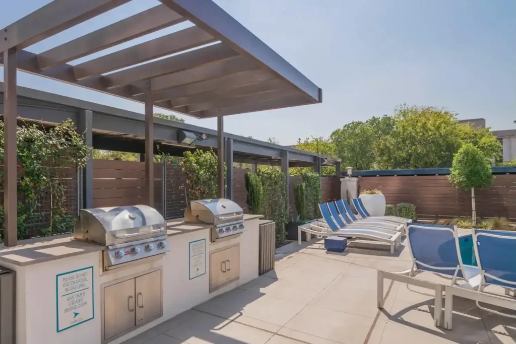 stainless steel grills and a seating area with blue lounge chairs, set on a sunny pool deck.
