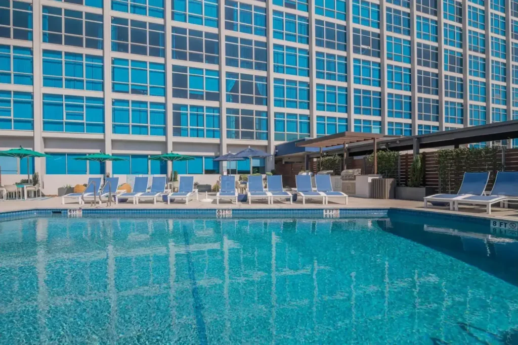 Swimming pool in a modern high-rise building, with clear water, white and blue lounge chairs, green and blue umbrellas, and a blue sky.