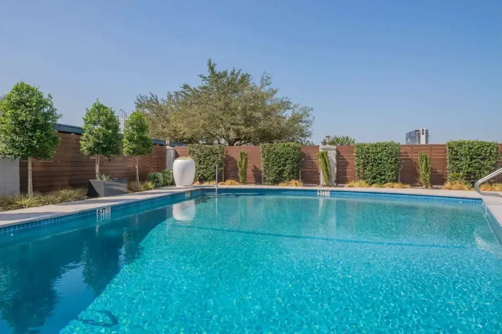 “Backyard with a rectangular swimming pool surrounded by a deck, potted plants, and a city skyline in the background.