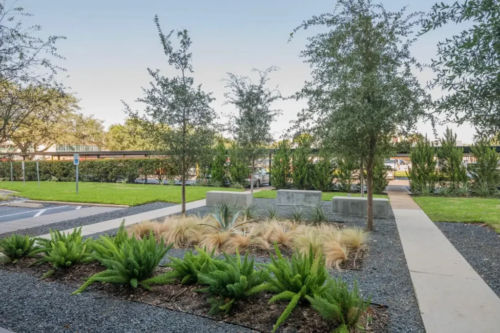 Suburban garden with modern design, featuring greenery, concrete planters, and a gravel pathway under a blue sky.