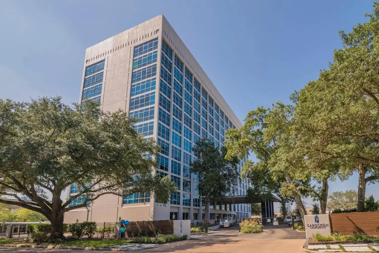 Entrance of Cabochon Apartments, a modern high-rise building with blue-tinted windows, landscaped with trees, shrubs, and grasses.