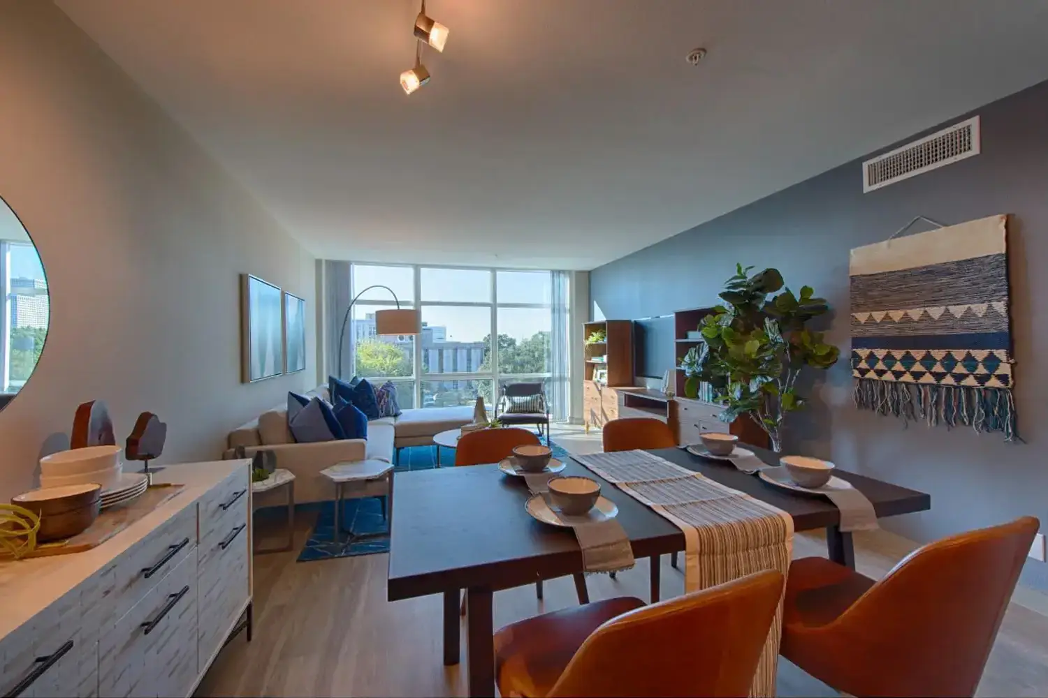 Modern living room with grey sofa, wooden coffee table, dining area with orange chairs, and city view window.