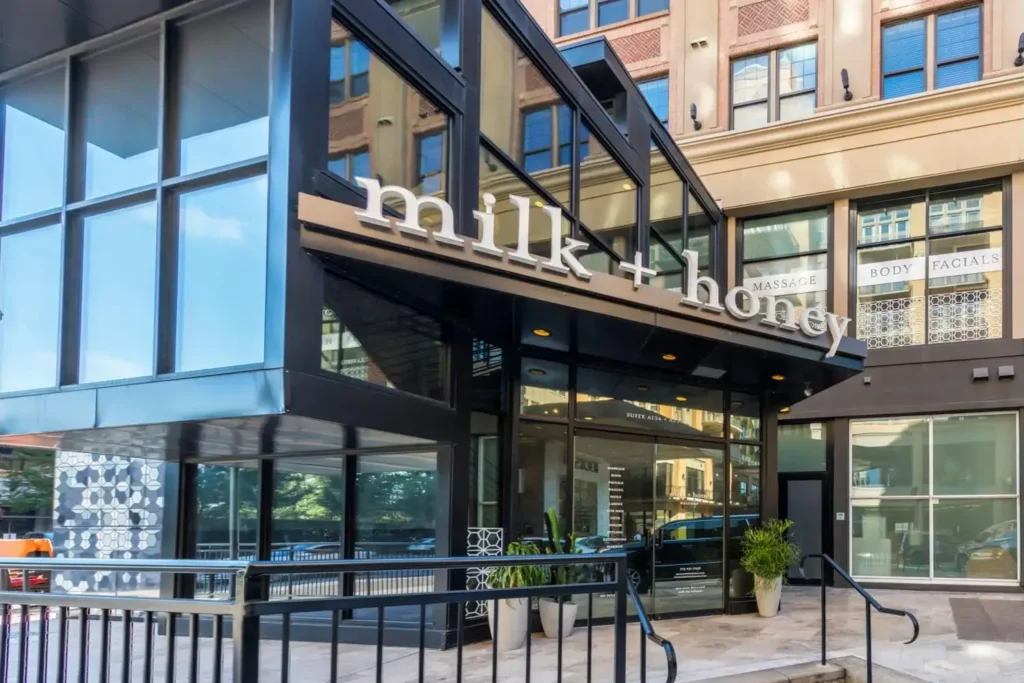 Modern building with a black metal awning and a sign reading ‘milk + honey’, surrounded by buildings.
