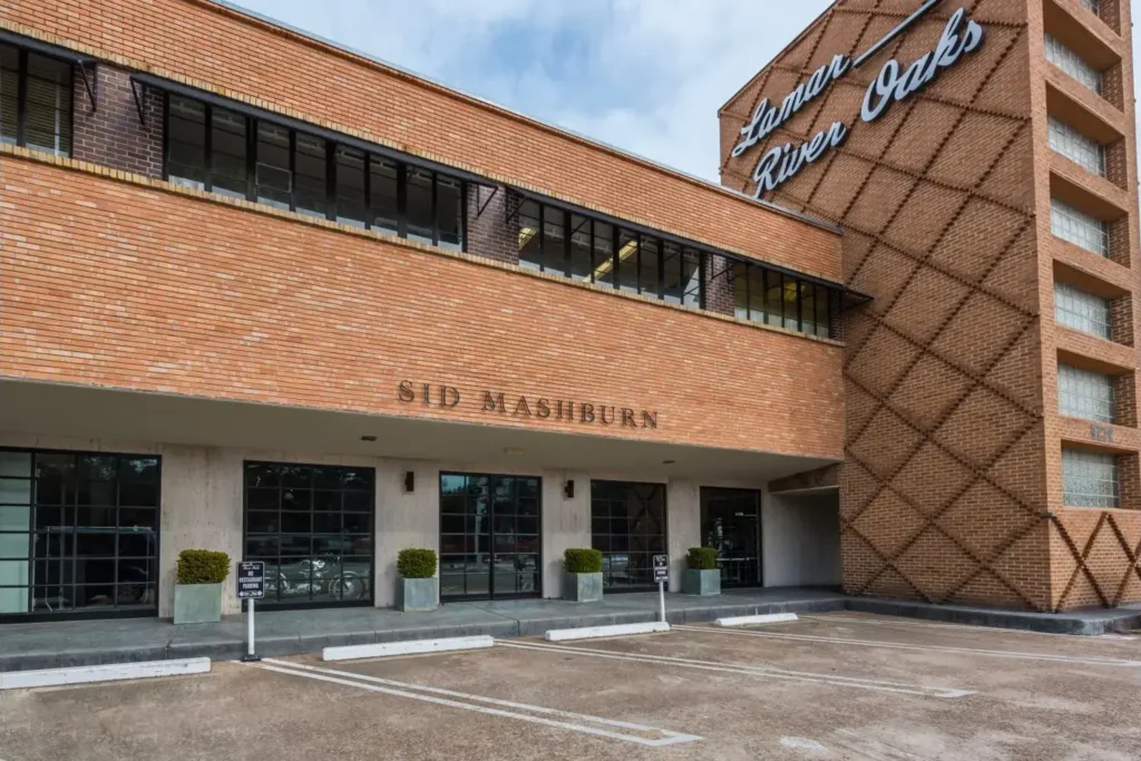 Brick building with a large glass window, a sign reading ‘Lamar River Oaks’, and an entrance flanked by potted plants.