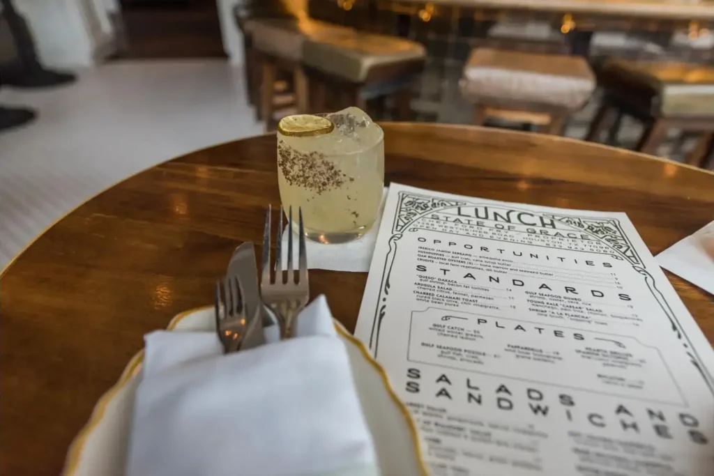 Restaurant table with a menu, a drink in a short glass, and a napkin with cutlery, set against a restaurant backdrop.