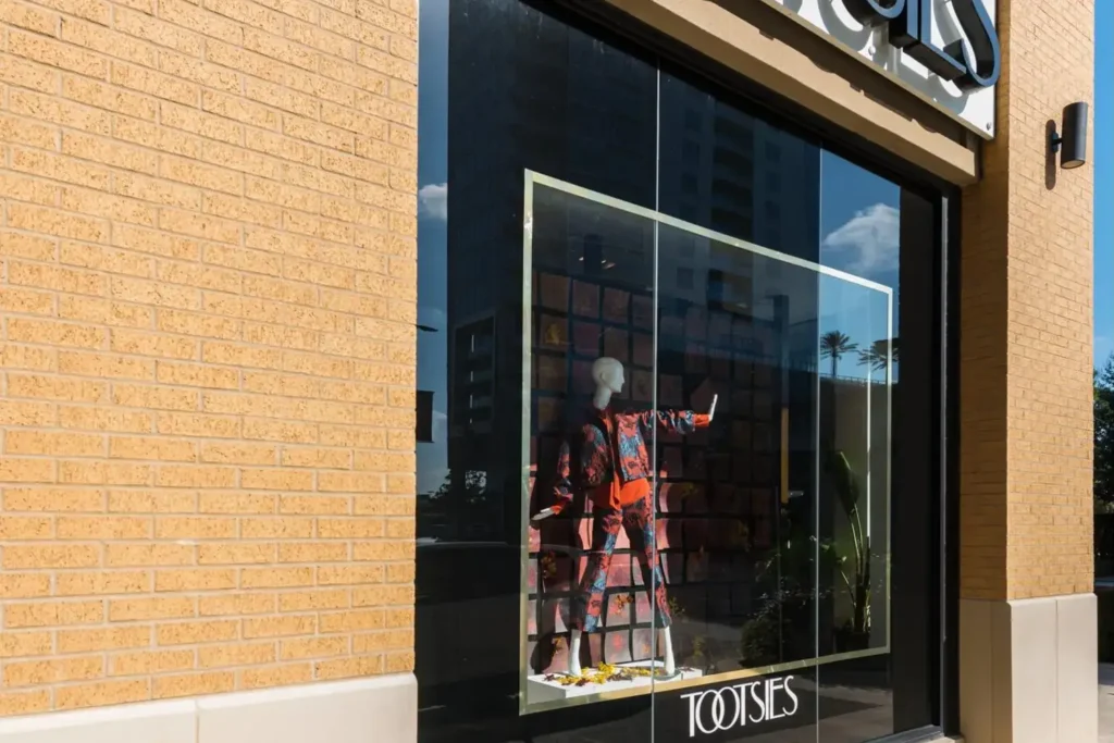 Storefront of ‘Tootsies’ with a window display featuring a mannequin in a red and black outfit, against a brick wall backdrop.