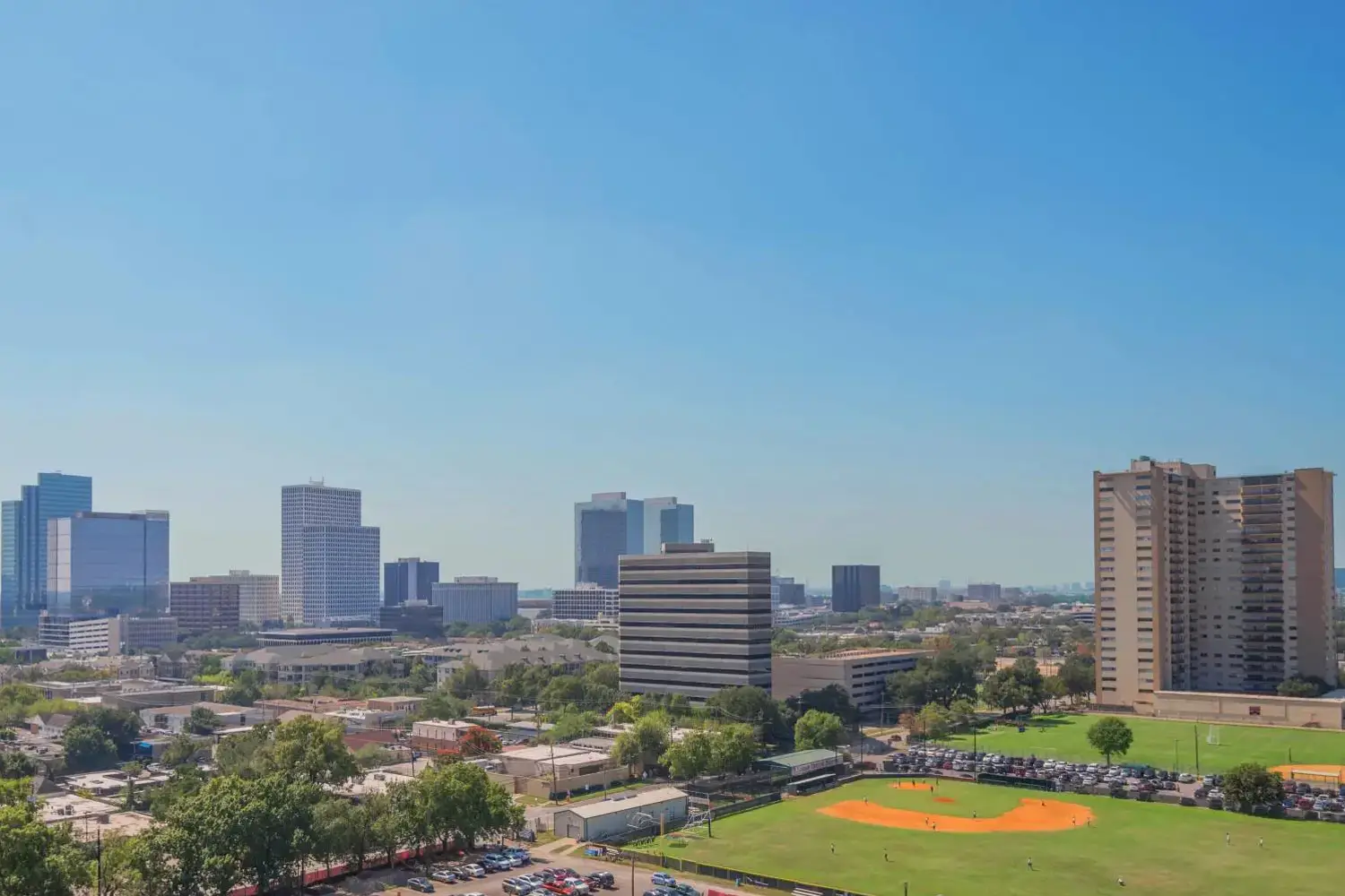 City skyline with a park in the foreground, featuring a large orange circle on the grass, under a clear blue sky.