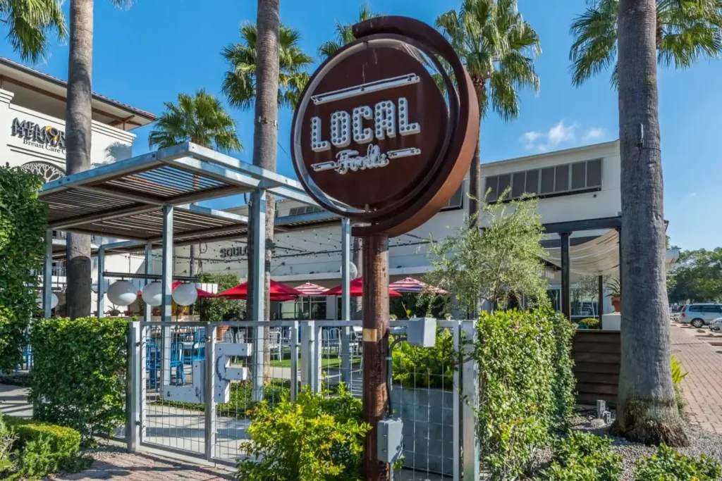 Outdoor seating area of a restaurant, with a metal pergola and red umbrellas, located in a shopping center.