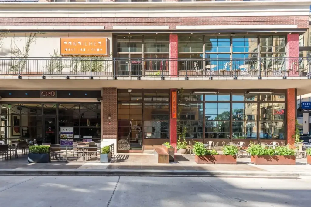 Modern storefront with a balcony, featuring a patio with tables and chairs, set against a backdrop of trees and a blue sky.