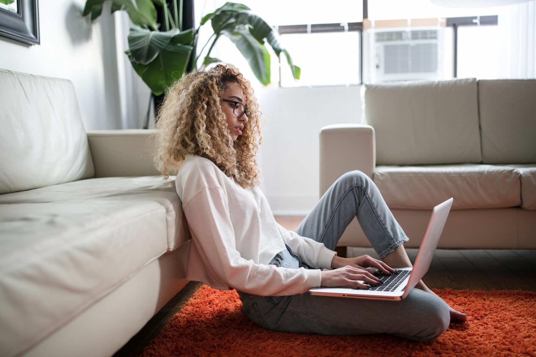 A person with curly hair sits on an orange rug, leaning against a white sofa while using a laptop. They are in a bright living room with large windows, plants, and light-colored furniture.